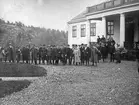 Samling herrar i hatt och en del i uniform poserar vid grusplan framför vitt hus med pelare. Centralen på utmarch 1911.