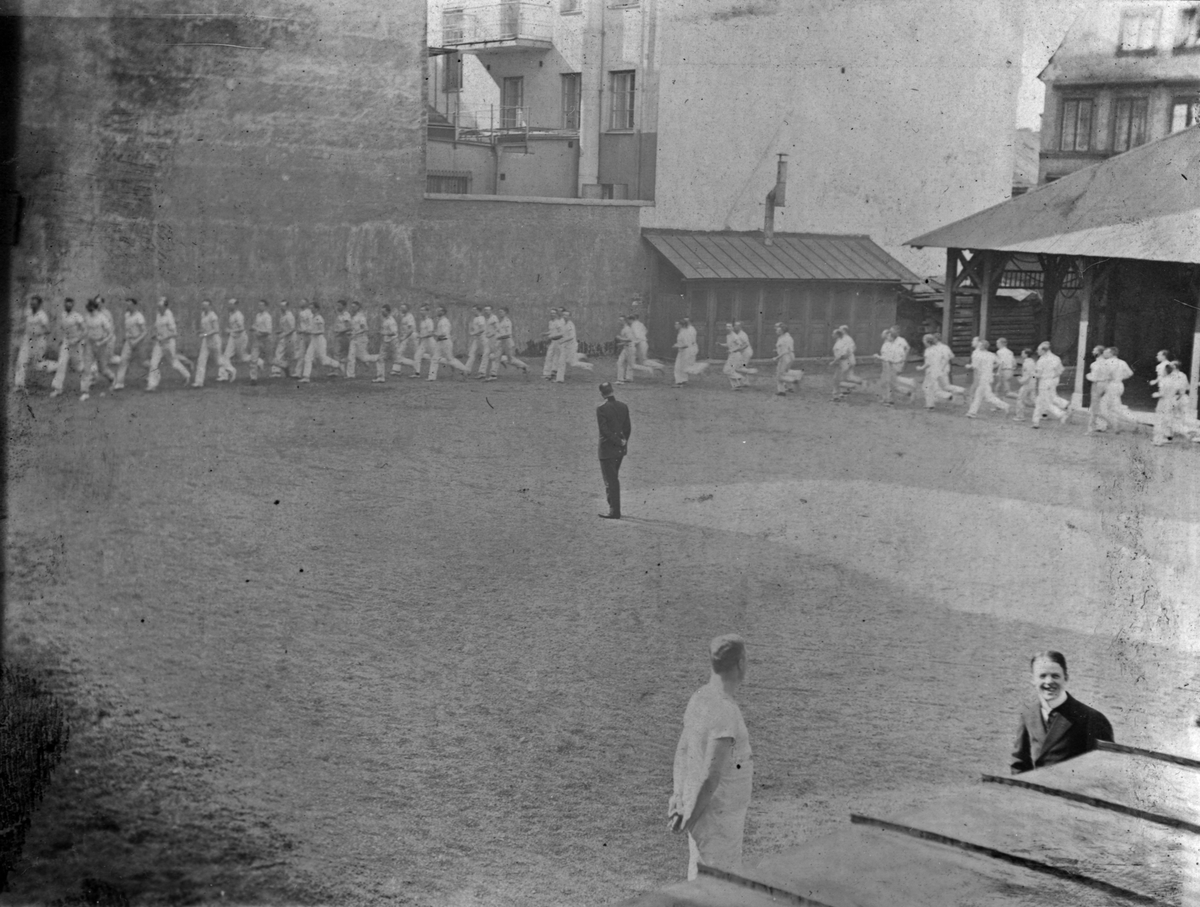 En samling män iklädda träningskläder springer på led om två runt en grusplan vid Gymnastiska central institutet (GCJ 1911-1912. I förgrunden syns två män varav en ler glatt mot kameran.
