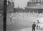 Gymnastiska central institutet. Män utövar gymnastik på gårdplan omgiven av hus under 1911-1912, alla klädda i vita träningskläder. Bredvid står tre män och inspekterar.