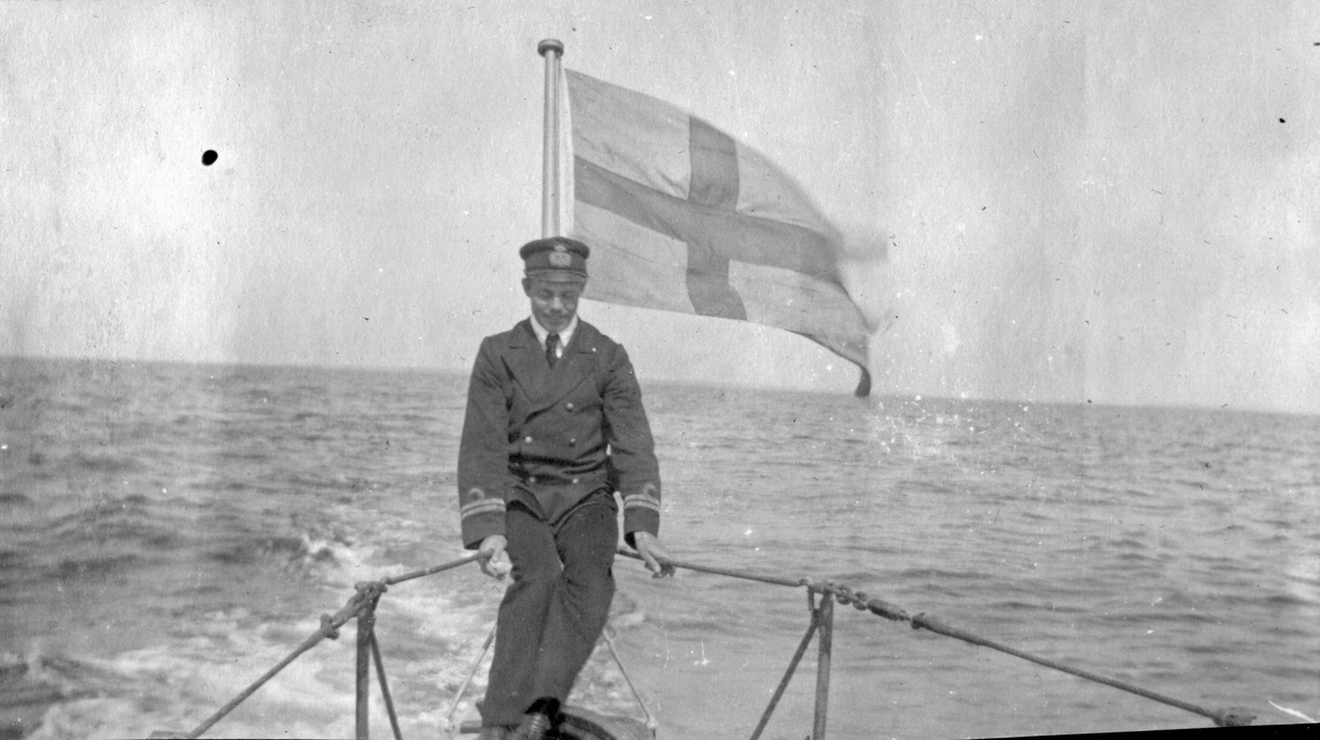 Sjöbefäl ombord på ubåt till sjöss som sitter på relingen. En flagga vajar i vinden bakom honom.