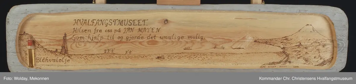 Haugenstranda med Beerenberg i bakgrunnen og to små båter med hvalbåt på slep mot Brielletårnet, mens fem mann venter på land ned flenseknivene klare.
