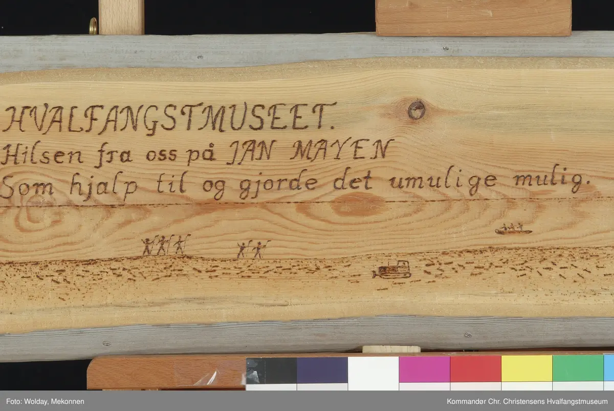 Haugenstranda med Beerenberg i bakgrunnen og to små båter med hvalbåt på slep mot Brielletårnet, mens fem mann venter på land ned flenseknivene klare.