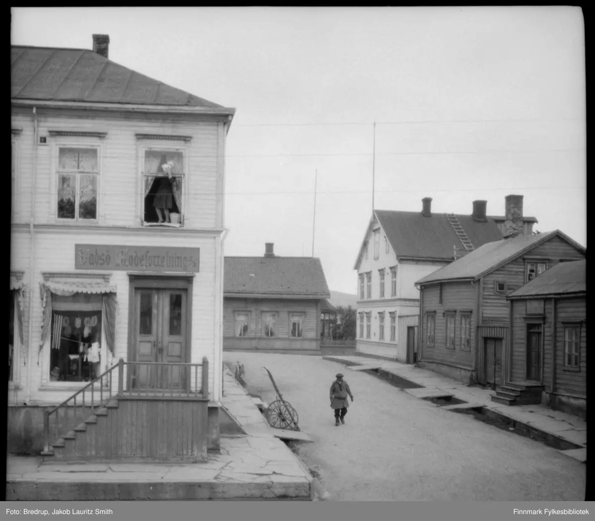 Vadsø sentrum rundt 1920-1930. En mann i kofte går forbi "Vadsø Modeforretning" drevet av søstrene Sundelin fra til krigen.  Det var vanlig å bruke kofte på denne tiden. Mannen kan være en same, men like godt han kan være en kven. En kvinne står i andre etasje og pusser vinduer. Det står en sykkel og ei kjerre parkert utenfor butikken.