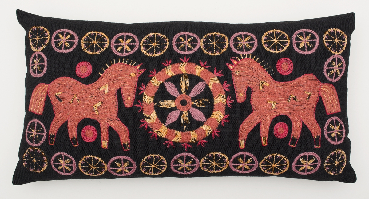 Stoppad kudde. Broderad på mörkbrunt broderulltyg från Klippan, färg 6. Broderad med Klippans broderiull- och lingarn i rött, orange och brunrosa. Två hästar som står vända mot varandra med ett hjul mellan dem. En blomma i hjulet. Små hjul i ram runt hästarna. Baksida i samma tyg.