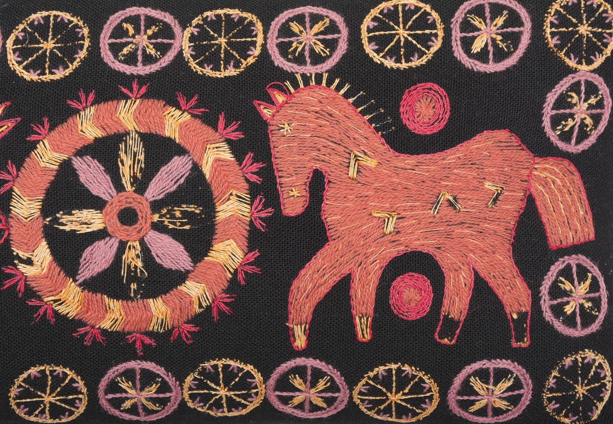 Stoppad kudde. Broderad på mörkbrunt broderulltyg från Klippan, färg 6. Broderad med Klippans broderiull- och lingarn i rött, orange och brunrosa. Två hästar som står vända mot varandra med ett hjul mellan dem. En blomma i hjulet. Små hjul i ram runt hästarna. Baksida i samma tyg.