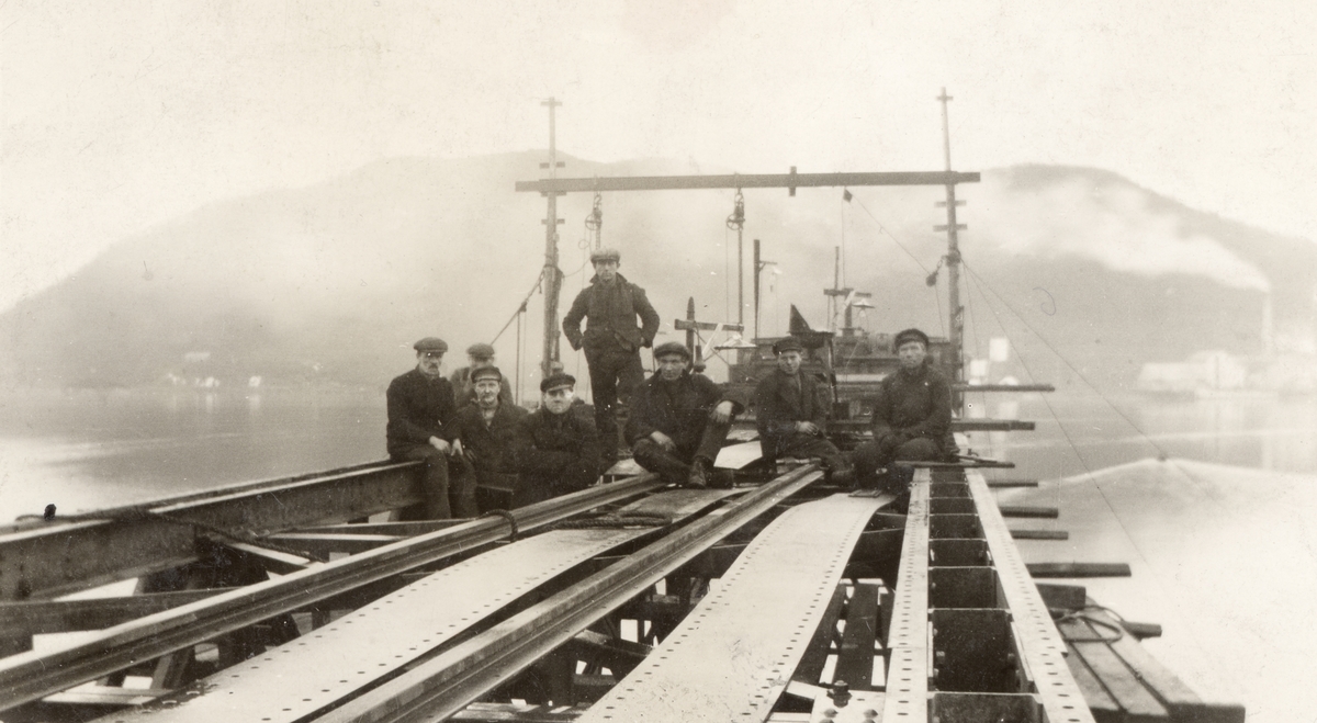 Gruppebilde av arbeidere, tatt i forbindelse med bygging av kullkranen hos B.J.N. Mercur Sildoljefabrikk i bakgrunnen til høyre.