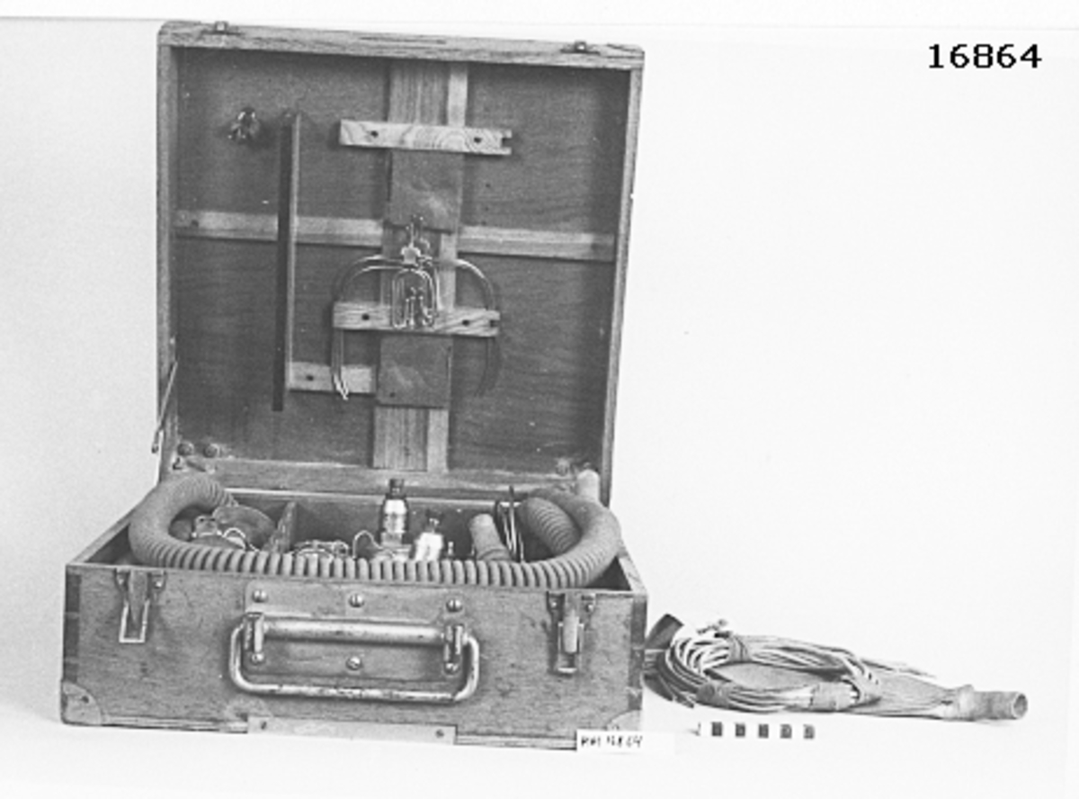 Låda av fernissat trä, försedd med lock på gångjärn och låsöverfall samt rörligt handtag i vitmetall på framsidan. Metallförstärkta hörn. Inuti lådan finns följande föremål: 2 st manometrar, 4 st andningsslangar textilklädd gummislang (L= 125 mm D= 33 mm samt 2 st små: L= 210 mm). 4 st munstycke för upphängning, 4 st metallföremål för upphängning, 2 st tredelade kopplingsrör i metall, 1 st metallbygel med skruv, 2 st byglar för hålla mask mot ansikte, 1 st nyckel passande till tre muttrar av olika storlek, 6 st gummislang (L= 1500 mm D= 4 mm), 1 st gummilunga.