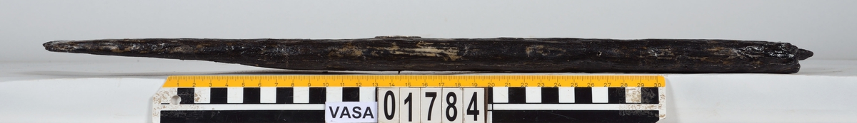 Möjligen en del av ett bord till en mindre båt. Den ena änden är avbruten, den andra utgörs av en bladlask med lång avfasning. Bevarad längd på fasningen är 130 mm men kan ha varit något längre ty kanten är avbruten. Bladlaskens fasning är  avgränsad av en tydlig ristad linje som bör har visat båtbyggaren var lasken skall börja. Den ena långsidan kan vara nära sin ursprungliga fason. Här finns en svag skålad ränna som kan vara en drevränna. I denna sitter ett bevarat spikhål. Ytterligare tre spikhål finns i bladlasken. Spikhålen är fyrkantiga. Den andra långsidan kan vara avbruten. Detta är svårt att säga säkert då ytan till största delen är anfrätt. Verktygsspår, sannolikt efter yxa, syns på laskens rätt så välbevarade yta.