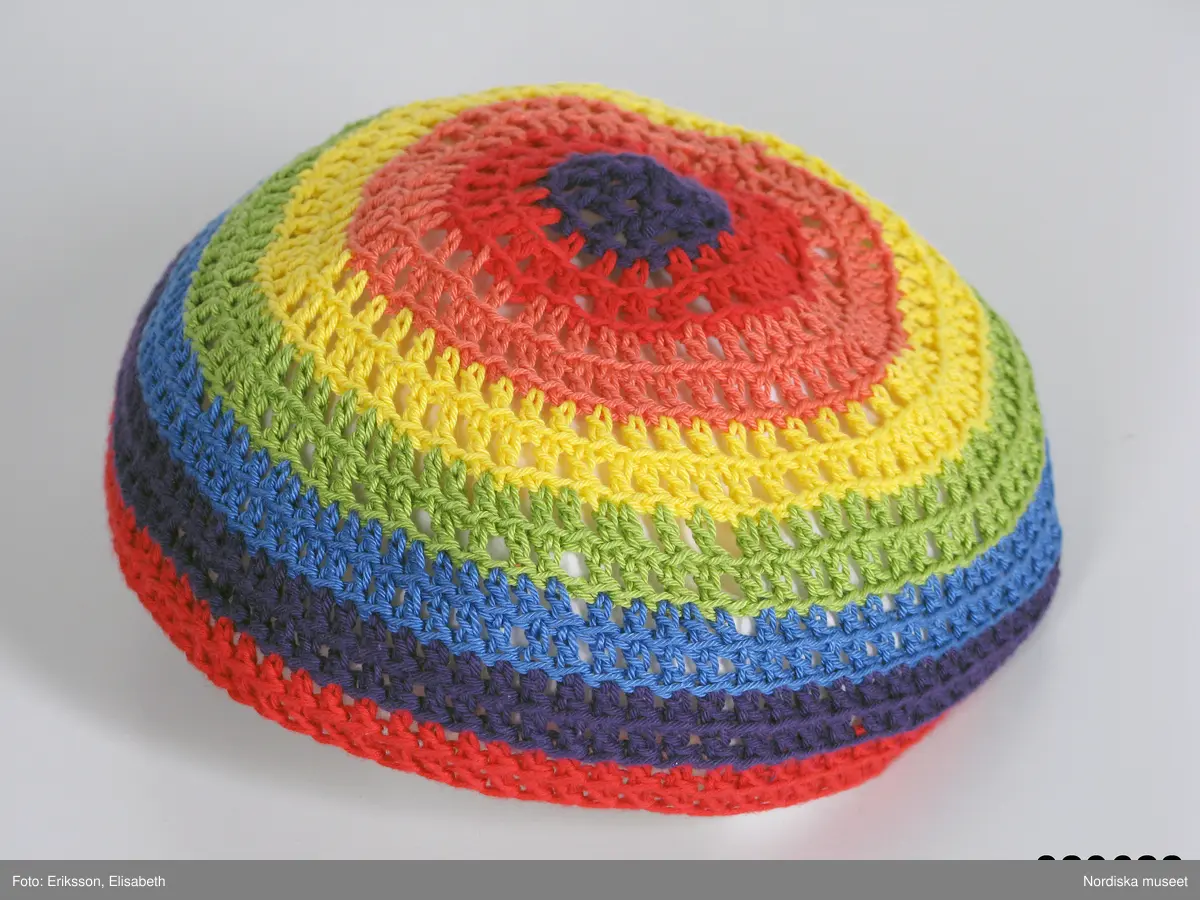 Kalott, glest virkad av bomullsgarn i randning (uppifrån och ner) lila, röd, rosa, gul, grön, blå, lila och röd. 
Matchande halsduk; se inv.nr 329.623. 
/Leif Wallin 2009-02-27