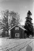Ett litet hus ligger bredvid stora träd, i förgrunden ett snöigt fält.