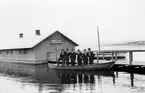 Sex män i en båt vid bryggan till Västra klapphuset vid Vätterstranden. Jönköping syns i bakgrunden.