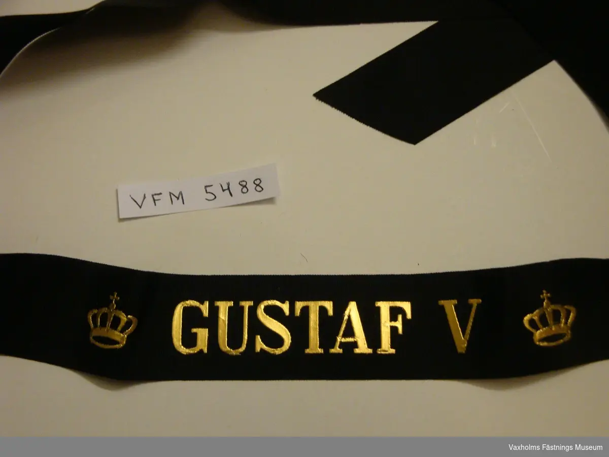 Mössband i svart sidenrips med guldtryck, kronor på var sida om texten; "GUSTAV V".