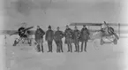Grupporträtt. Sex stycken militära flygare och två hundar framför flygplan Thulin K och Triplanet på flygfältet på Flygkompaniet på Malmen, vintertid, 1918. Från vänster: Gösta D:son Carlson, Gustaf von Segebaden, Nils Rodéhn, Paul af Uhr, flygspanare B. T. Kjelldahl, Fredrik Adilz.