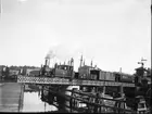 Ett tåg på UVHJ-banan passerar den gamla svängbron i anslutning till järnvägsstationen i Vänersborg.
Foto fr. ca 1915.
