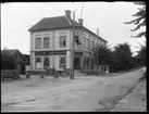 Olof Jonssons text: Charls Anderssons och Gustaf Svenssons hus i Strömslund. 1910 så bodde här 15 personer bl a Handlande Gustaf Adolf Svensson, Väne-Ryr 1881, hans hustru Linnea Ekman, Stora Tuna 1887 och deras dotter Birgit Linnea, Vassända-Naglum 1910. Gissningsvis kan det vara herr och fru Svensson som står på balkongen ovanför affären - som ägare till fastigheten borde de ju bo i lägenheten med balkong.