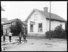 Fastigheten med två hus tillhörande f d poliskonstapel Johan Alfred Skogsbergs, Västra Tunhem 1858 och hans hushåll. 1910 så bodde han här tillsammans med sin hustru Hulda Sofia Spång, Naglum 1859, dottern Alfrida Kristina, Flakeberg 1892 och sonen Torsten Emanuel, Vassända-Naglum 1897. I huset till vänster på bilden fanns ett Nykterhetskafe och huset till höger har skylten E A Kihlbergs Speceriaffär. Barnen på gatan är okända.