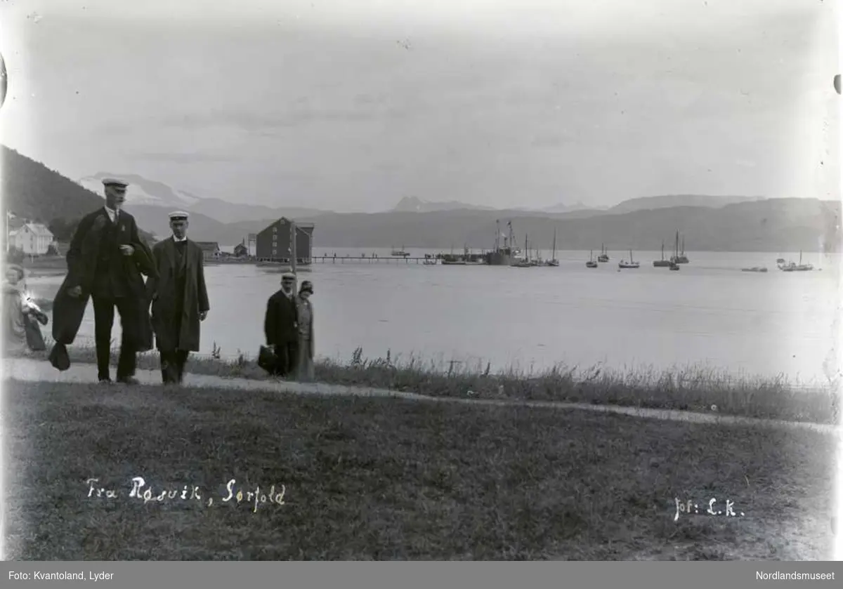 Folk trolig på tur fra brygga til kirka i forgrunnen. Brygga og vågen i Røsvik fotografert mot nord.
