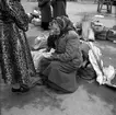 Denna bild är troligen från Stockholm dit fotograf Gustav Andersson ibland reste. På ett torg en sitter en äldre kvinna omgiven av väskor och påsar, en med texten 