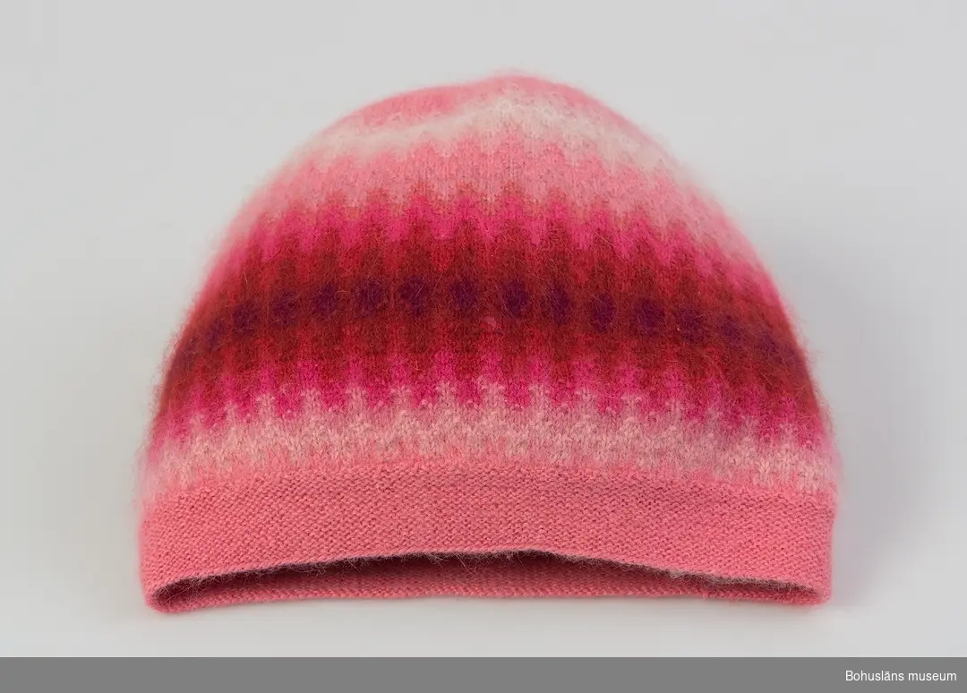 Stickad mössa i sotarmodell med rätstickad nederkant. Mössan är mönsterstickat med räta och aviga maskor i fyra olika rosa färger, cerise, tegelfärg, ljusbrunt, blått, beige och vitt mot en rosa botten. Ett mörkt löstagbart innerstöd av syntetmaterial finns. Jfr uppgifter på UM027468.