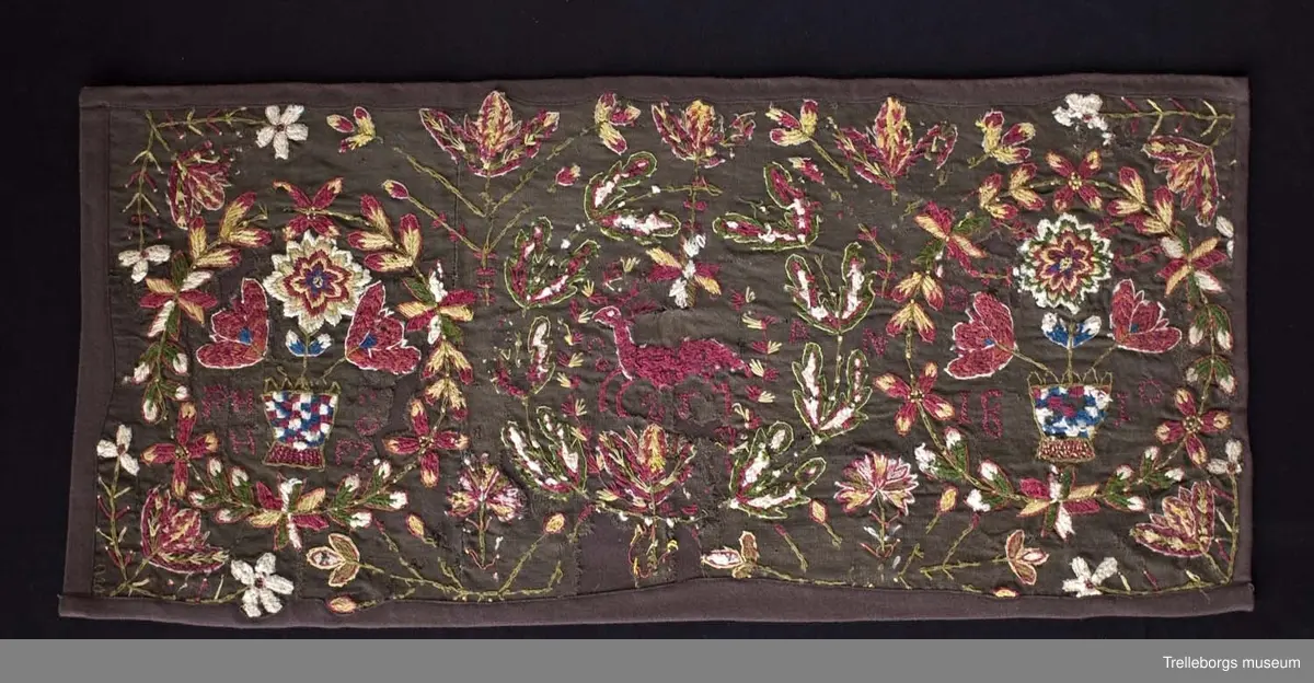 Broderad agedyna på mörkbrun botten. Motivet är tre stora blomkransar, de två yttersta har varsin rutig blomvas med tre blommor i, samt bokstäver PHH  OPZ, i den första och årtalet 1890 i den sista. I den mittersta kransen springer en stor röd hjort med röda horn. Även här finns bokstäver, två bakvända S och ett T. Färgerna går i rött, gult, grönt, vitt och blått. Utanför blomsterkransarna fortsätter blommor att växa upp.