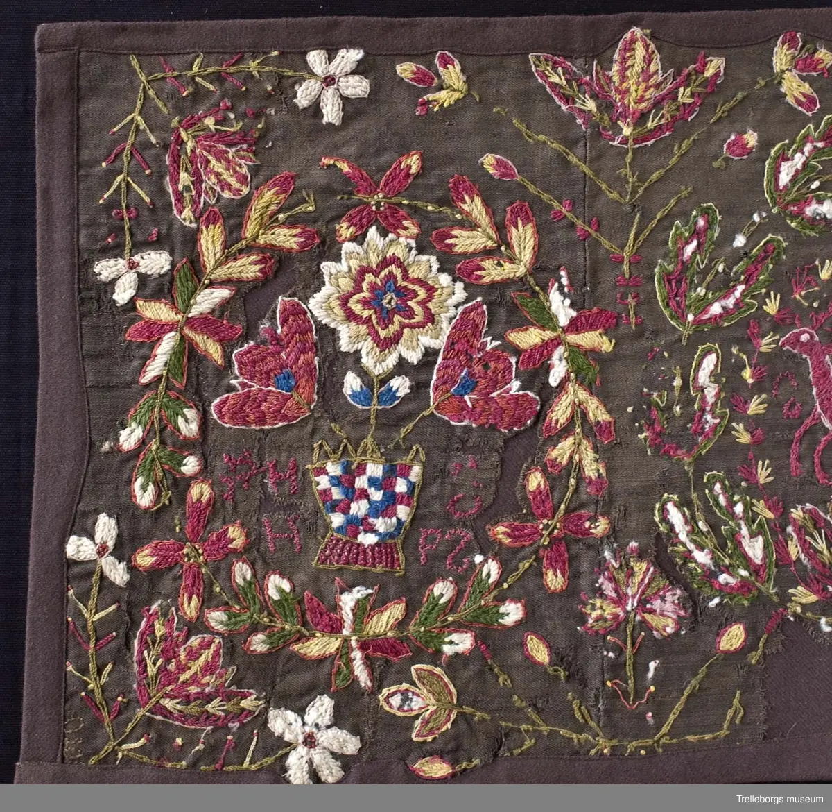 Broderad agedyna på mörkbrun botten. Motivet är tre stora blomkransar, de två yttersta har varsin rutig blomvas med tre blommor i, samt bokstäver PHH  OPZ, i den första och årtalet 1890 i den sista. I den mittersta kransen springer en stor röd hjort med röda horn. Även här finns bokstäver, två bakvända S och ett T. Färgerna går i rött, gult, grönt, vitt och blått. Utanför blomsterkransarna fortsätter blommor att växa upp.