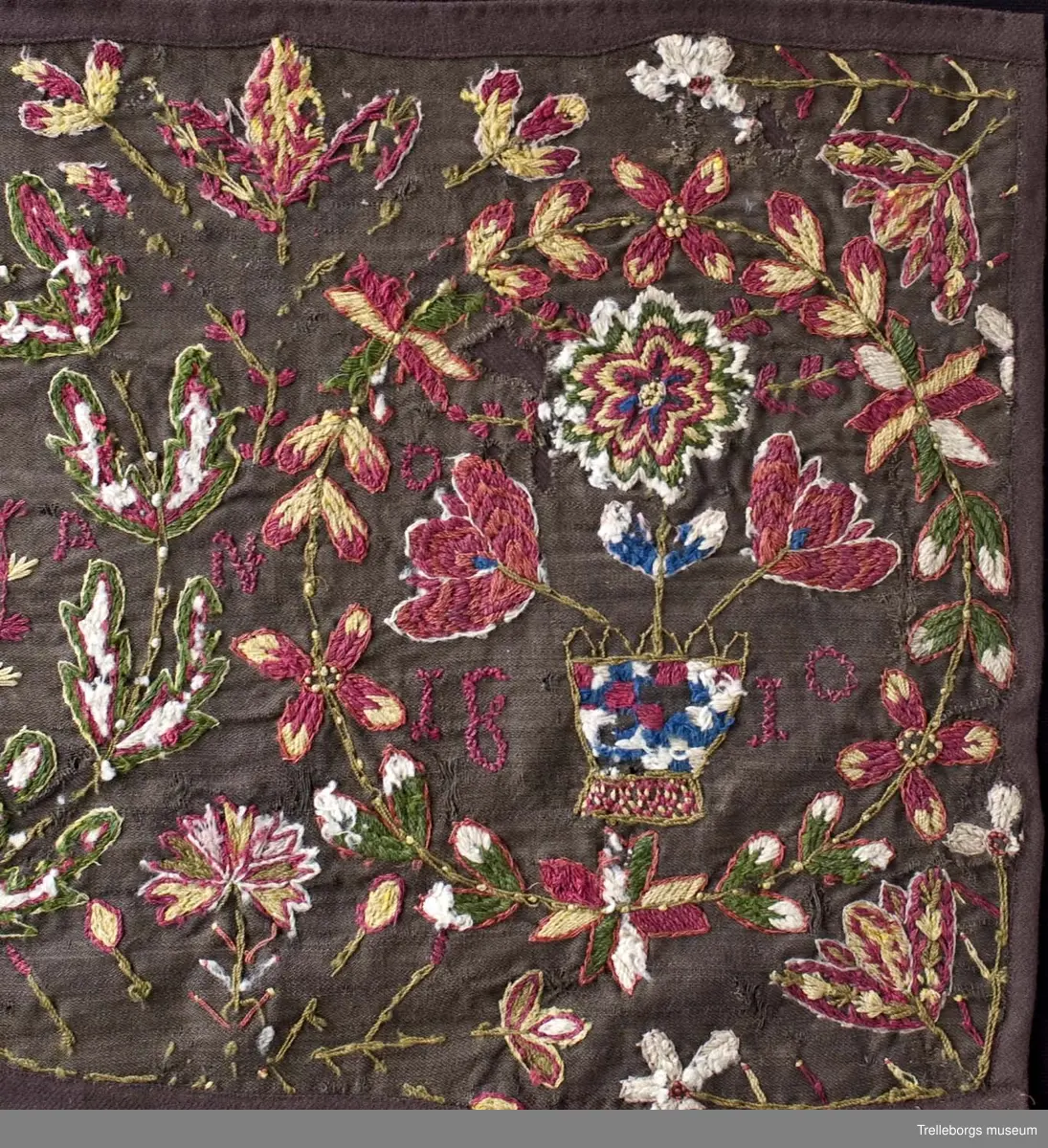 Broderad agedyna på mörkbrun botten. Motivet är tre stora blomkransar, de två yttersta har varsin rutig blomvas med tre blommor i, samt bokstäver PHH  OPZ, i den första och årtalet 1890 i den sista. I den mittersta kransen springer en stor röd hjort med röda horn. Även här finns bokstäver, två bakvända S och ett T. Färgerna går i rött, gult, grönt, vitt och blått. Utanför blomsterkransarna fortsätter blommor att växa upp.