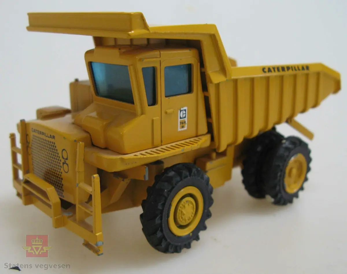 Leketøy, miniatyr tipptruck, Caterpillar 769 B, av metall, gummi og plexiglass. Gul med svarte hjul. Maskina har to akslinger, (seks hjul), og lastekasse som kan tippes opp. Merking fra produsenten under maskina samt Caterpillar merking utenpå.