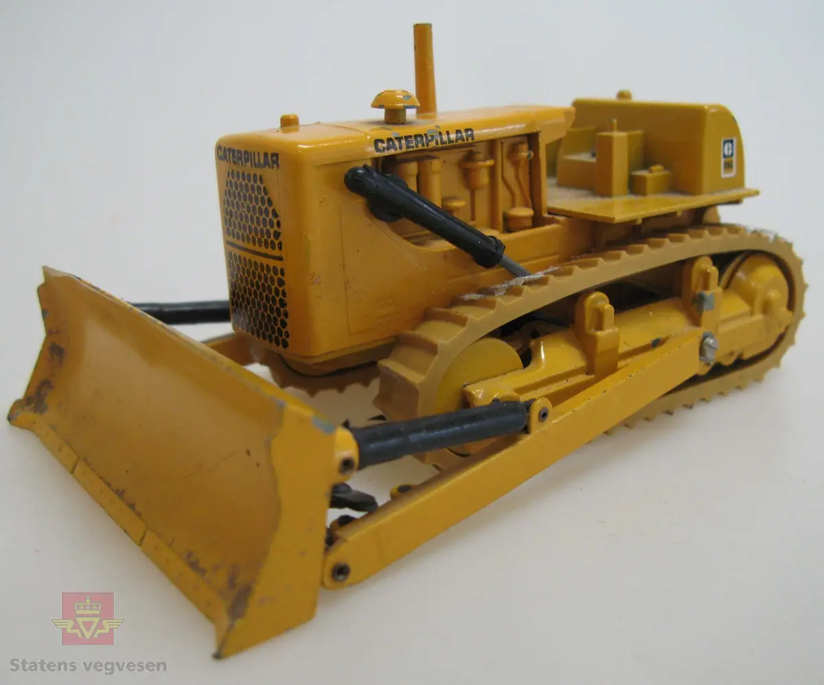 Leketøy, miniatyr bulldozer, Caterpillar D9G, av metall og gummi. Gul med enkelte svarte detaljer. Dozeren går på belter, har skjær foran og trekkfeste bak. Merking fra produsenten under leketøyet samt Caterpillar merking utenpå.