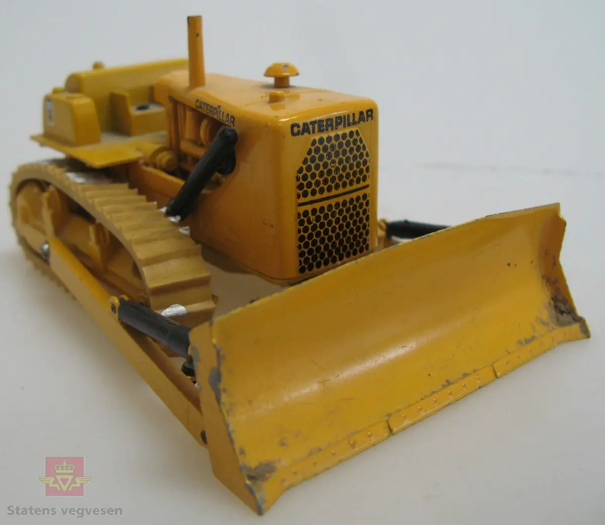 Leketøy, miniatyr bulldozer, Caterpillar D9G, av metall og gummi. Gul med enkelte svarte detaljer. Dozeren går på belter, har skjær foran og trekkfeste bak. Merking fra produsenten under leketøyet samt Caterpillar merking utenpå.