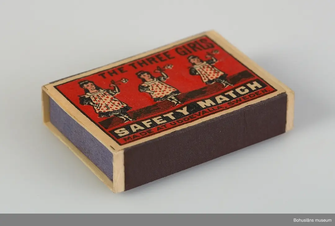 En tändstickask med teckning av tre flickor i förkläden och med texten:
THE THREE GIRLS
SAFETY MATCH
MADE IN UDDEVALLA, SWEDEN