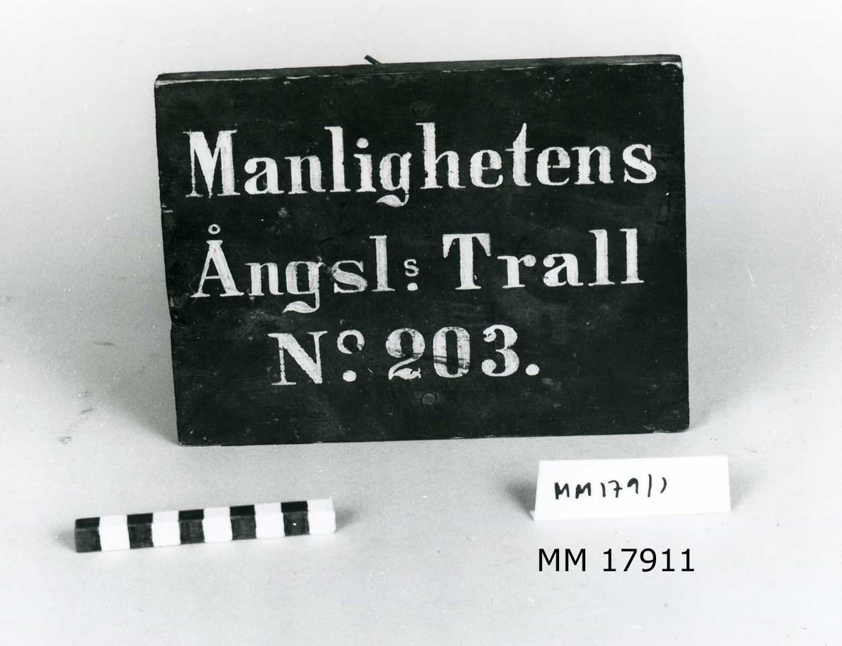 Skylt av trä, svartmålad framsida med målade bokstäver i vitt, tre rader: " Manlighetens Ångsls. Trall N 203 . " På baksidan samma text, här med blå krita.