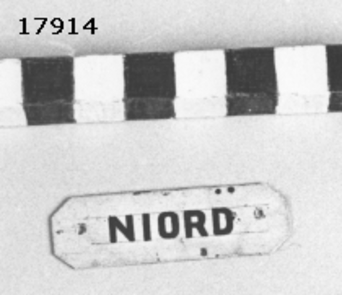 Rektangulär skylt av plåt med avklippta hörn. Framsidan vitmålad och försedd med handmålad text i svart: " NIORD ". Hål i kanterna för spikar.
