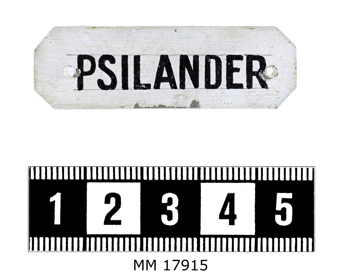 Rektangulär skylt av plåt med avklippta hörn. Framsidan vitmålad och försedd med handmålad text i svart: " PSILANDER ". Hål i kanterna för spikar.