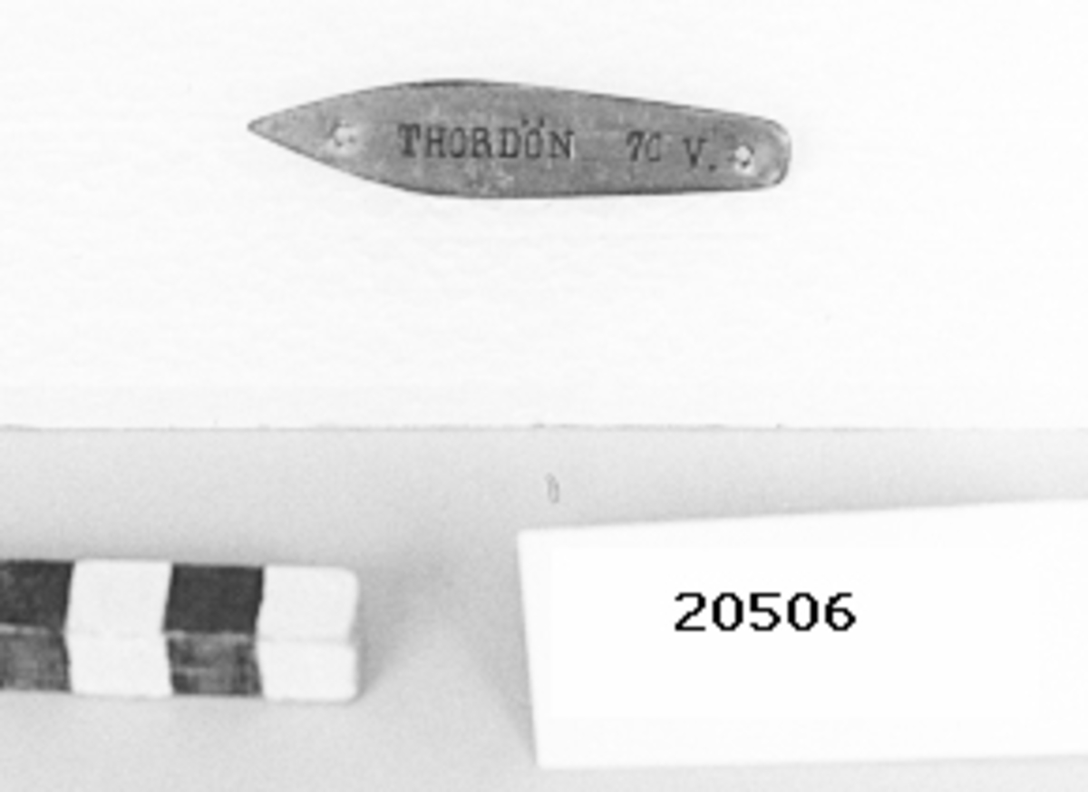 Skylt av mässingsplåt, i formen som ett fartyg med hål i för och akter för upphängning. Instansad text på mitten: "THORDÖN 70 V.".