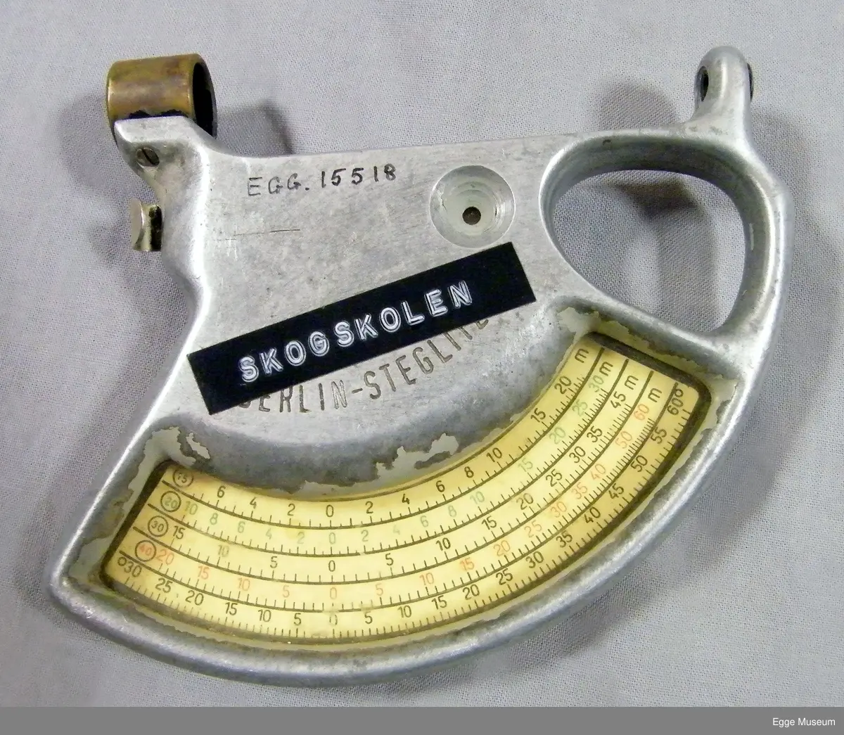 Høyemåler i lettmetal (aluminium?) med rektangulær skulderveske. Formen på instrumentet er omtrent en kvart sirkel. Instrumentet har gripehull i bakkant. Over gripehull er det montert et siktehull. I fremre kant er det montert en messingring, inne i denne ringen er det to spisse og triangulære spisser som stikker ut og peker mot hverandre. På ene siden av instrument er det montert et metallskilt med beregningsskala (ant tabellgrenser for avvik ved forskjellige målinger). På den andre siden er det en måleskale/avlesningsskala med en bevegelig pendel/fjær, dekt med gjenomsiktig plast.

Instrumentet er oppbevart i ei bæreveske av plasttrukket, grønt lerretsstoff. Veska har skulderreim i lær, som er poppet fast til vesken sider. Veska lukkes med lokk og låsestropp (denne er avslitt). Veska har to rom innvendig. Veska har muggskader på baksiden, samt at ene hjørnet i bunn har skader som har blitt sydd sammen.

Høydemåler for bruk på stående skog. Når man siktet inn toppen (eller rota) på treet som stod mellom disse punktene, og samtidig trykket inn en fjær, kunne man lese av vinkel og høyde, sett i forhold til en horisontal linje fra siktepunktet, i en måleskala på instrumentet. Man skulle vanligvis stå 20 meter fra treet. Høydemåleren er grålakkert, med noe avflassing. Av den tyske fabrikaten Blume-Leiss.

Brukt i undervisninga ved Skogskolen på Steinkjer.