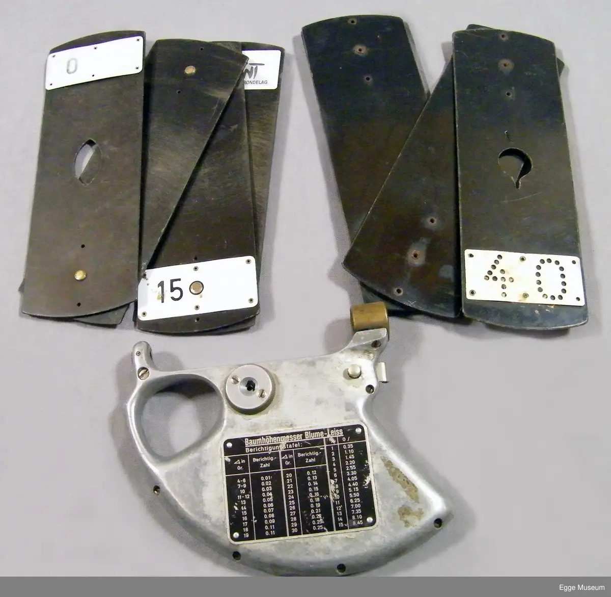 Høyemåler i lettmetal (aluminium?) med rektangulær skulderveske. Formen på instrumentet er omtrent en kvart sirkel. Instrumentet har gripehull i bakkant. Over gripehull er det montert et siktehull. I fremre kant er det montert en messingring, inne i denne ringen er det to spisse og triangulære spisser som stikker ut og peker mot hverandre. På ene siden av instrument er det montert et metallskilt med beregningsskala (ant tabellgrenser for avvik ved forskjellige målinger). På den andre siden er det en måleskale/avlesningsskala med en bevegelig pendel/fjær, dekt med gjenomsiktig plast.

Instrumentet er oppbevart i ei bæreveske av plasttrukket, grønt lerretsstoff. Veska har skulderreim i lær, som er poppet fast til vesken sider. Veska lukkes med lokk og låsestropp (denne er avslitt). Veska har to rom innvendig. Veska har muggskader på baksiden, samt at ene hjørnet i bunn har skader som har blitt sydd sammen.

Høydemåler for bruk på stående skog. Når man siktet inn toppen (eller rota) på treet som stod mellom disse punktene, og samtidig trykket inn en fjær, kunne man lese av vinkel og høyde, sett i forhold til en horisontal linje fra siktepunktet, i en måleskala på instrumentet. Man skulle vanligvis stå 20 meter fra treet. Høydemåleren er grålakkert, med noe avflassing. Av den tyske fabrikaten Blume-Leiss.

Brukt i undervisninga ved Skogskolen på Steinkjer.