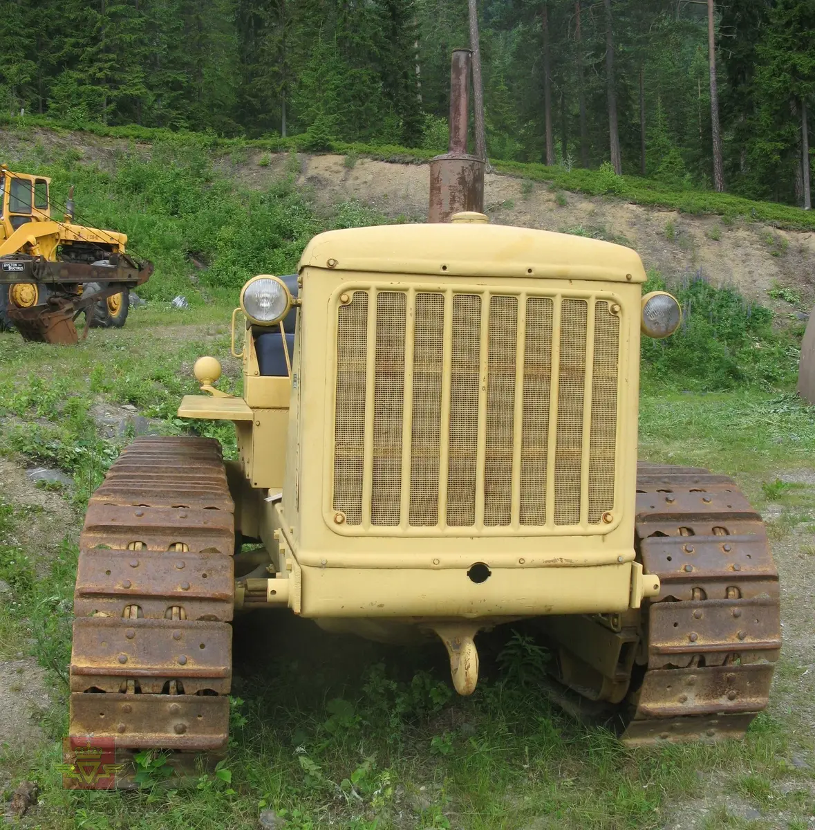 Caterpillar D 7. Gul, (opprinnelig grønn), med svart sete. Maskina er egentlig en bulldozer, men denne er bygget som beltetraktor. Forskjellen ser man best på den bredere fronten hvor det ikke er plasss til hydrauliksylindre for løft av bulldozerskjær. Denne har likevel hatt et skjær, men løfteanordningen som ble benyttet var vinsjen som satt bak på maskina. Wirene gikk fra vinsjen og over to tårn og videre ned på skjæret. Maskina har en 4-sylindret dieseldrevet motor som yter ca 128 Hk. Startmotoren er bensindrevet.
Oppstarting av dieselmotorene er såpass spesiell at den beskrives her:
Starteren er en 2-sylindret bensinmotor. Etter å ha skrudd opp bensinkrana og slått på tenningsbryteren på ON, settes sveiva ned gjennom panseret. Choken må setes på etter behov og gasspjeldet må settes på litt over tomgang. Startmotoren dras over komp. til den starter. Startmotoren bør gå noen minutter fordi avgassene varmer opp dieselmotorens innsugningsrør. Dieselmotorens gasshåndtak settes på halv gass, og dekompresjonshåndtaket på siden av motoren settes til, (slik at motoren går lett rundt).
Det finnes to håndtak for å koble til driften mellom starter og hovedmotor, det ene for å flytte "bendixdrevet" i inngrep med svinghjulet og det andre håndtaket er en clutch som setter i gang bevegelsen av dieselmotoren. Man kjører da noen omdreininger får så å sette dekompresjonshåndtaket i halvstilling. Når man ser på eksosen at dieselen begynner å forbrenne slår man håndtaket over på full kompresjon. Etter noen omdreininger går hovedmotoren av seg selv, slik at starteren kan kobles fri og bensinkrana stenges og tenning slåes av.