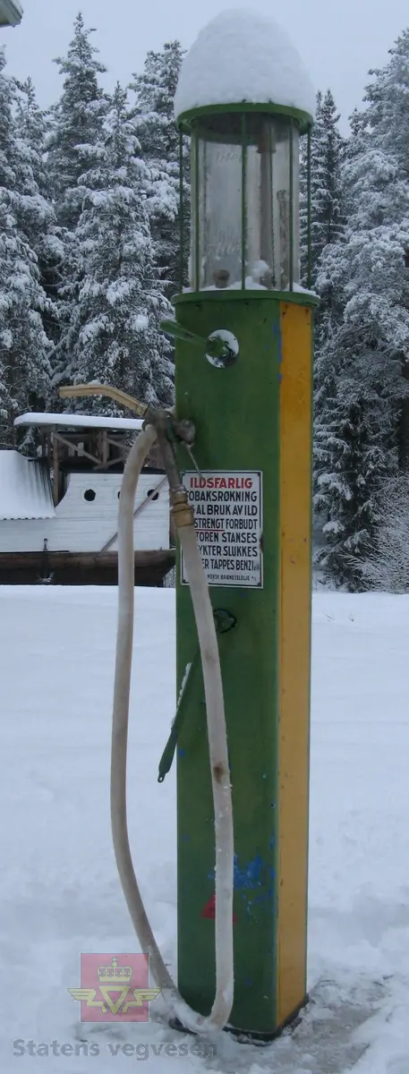 Flerfarget, hovedfarger er grønn og gul, (opprinnelig blå). Håndpumpe og porsjoneringsglass. Står på sokkel. Merking.
Trekasse med lokk høre til pumpa.