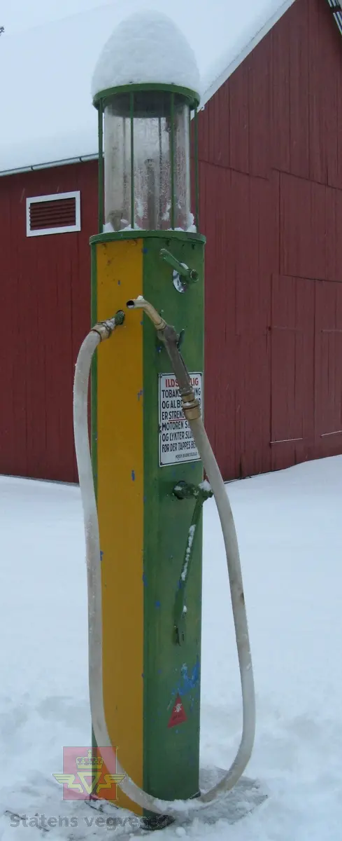 Flerfarget, hovedfarger er grønn og gul, (opprinnelig blå). Håndpumpe og porsjoneringsglass. Står på sokkel. Merking.
Trekasse med lokk høre til pumpa.