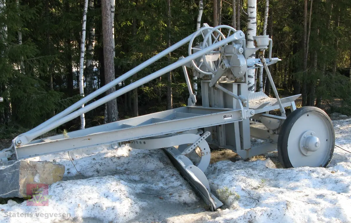 Grå veghøvel/skrape med 2 hjul, (enakslet). Dekk av kompakt gummi. Sete for fører. Ratt for justering av skjær. Har drag for å trekkes av traktor eller lastebil. 
Ligner noe på en Odin høvel.