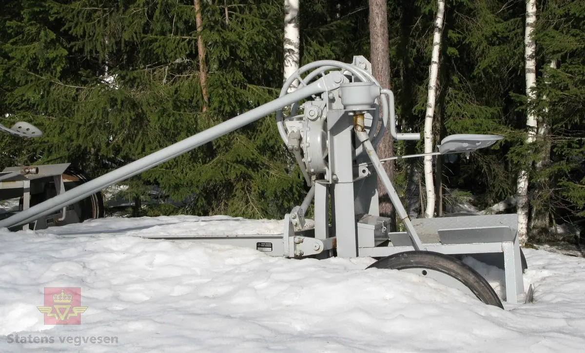 Grå veghøvel/skrape med 2 hjul, (enakslet). Dekk av kompakt gummi. Sete for fører. Ratt for justering av skjær. Har drag for å trekkes av traktor eller lastebil. 
Ligner noe på en Odin høvel.