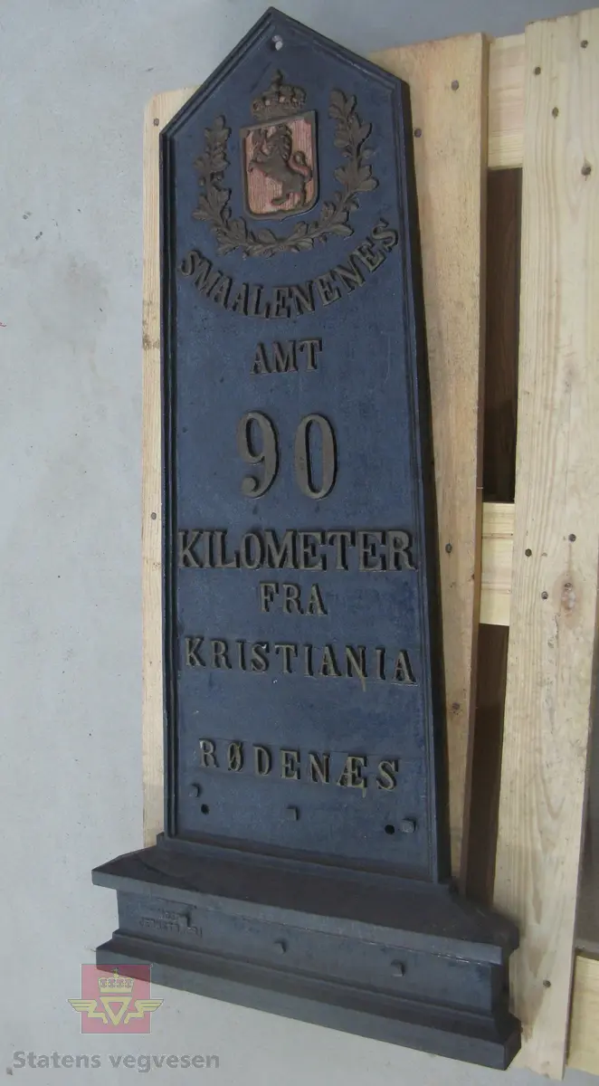 Kilometertavle, (skilt), lagd av støpejern, med riksvåpenmotiv og teksten:" SMAALENENES AMT 90 KILOMETER FRA KRISTIANIA RØDENÆS". Svart bunnfarge, tekst i gull. Baksiden har teksten: "MOSS JERNSTØBERI".