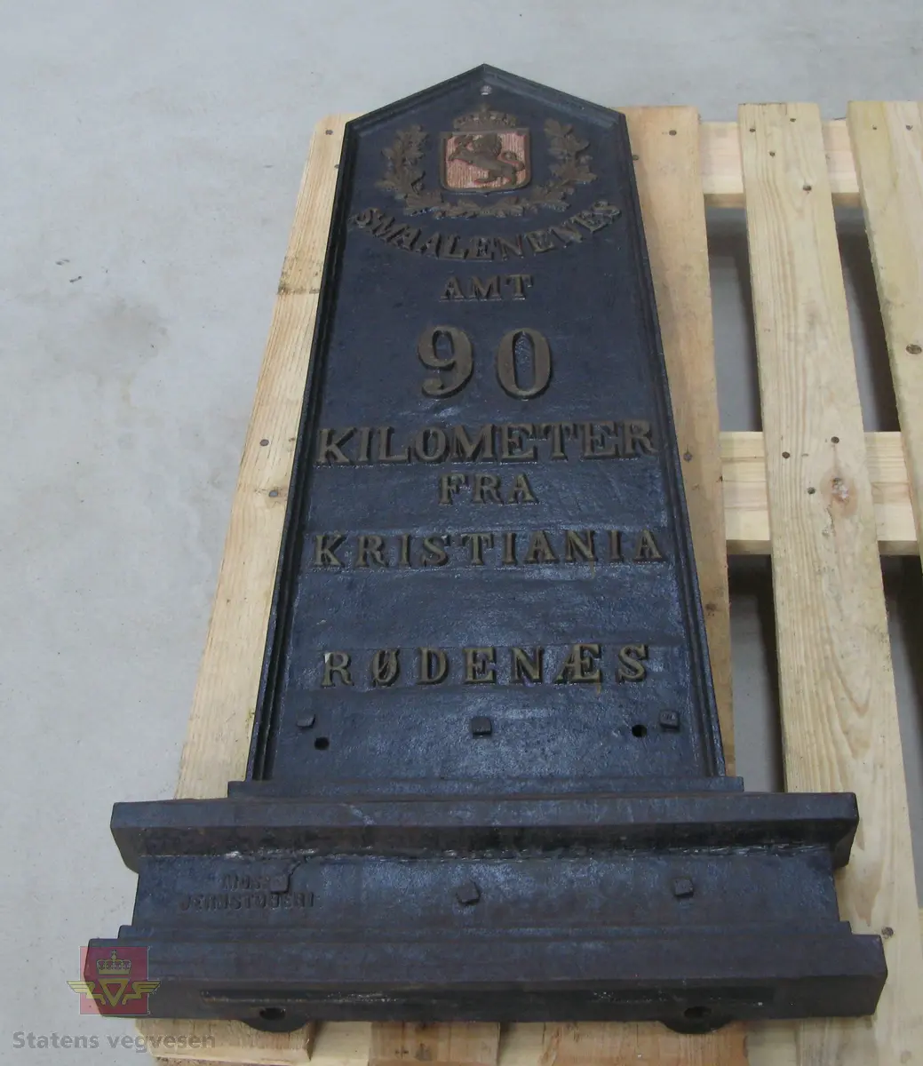 Kilometertavle, (skilt), lagd av støpejern, med riksvåpenmotiv og teksten:" SMAALENENES AMT 90 KILOMETER FRA KRISTIANIA RØDENÆS". Svart bunnfarge, tekst i gull. Baksiden har teksten: "MOSS JERNSTØBERI".
