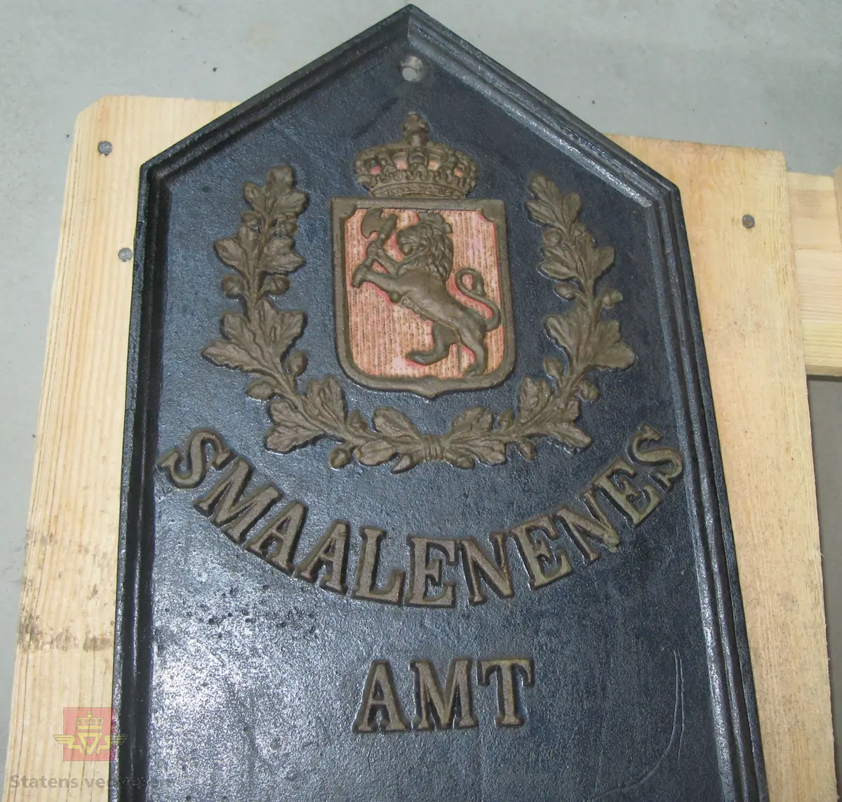 Kilometertavle, (skilt), lagd av støpejern, med riksvåpenmotiv og teksten:" SMAALENENES AMT 90 KILOMETER FRA KRISTIANIA RØDENÆS". Svart bunnfarge, tekst i gull. Baksiden har teksten: "MOSS JERNSTØBERI".