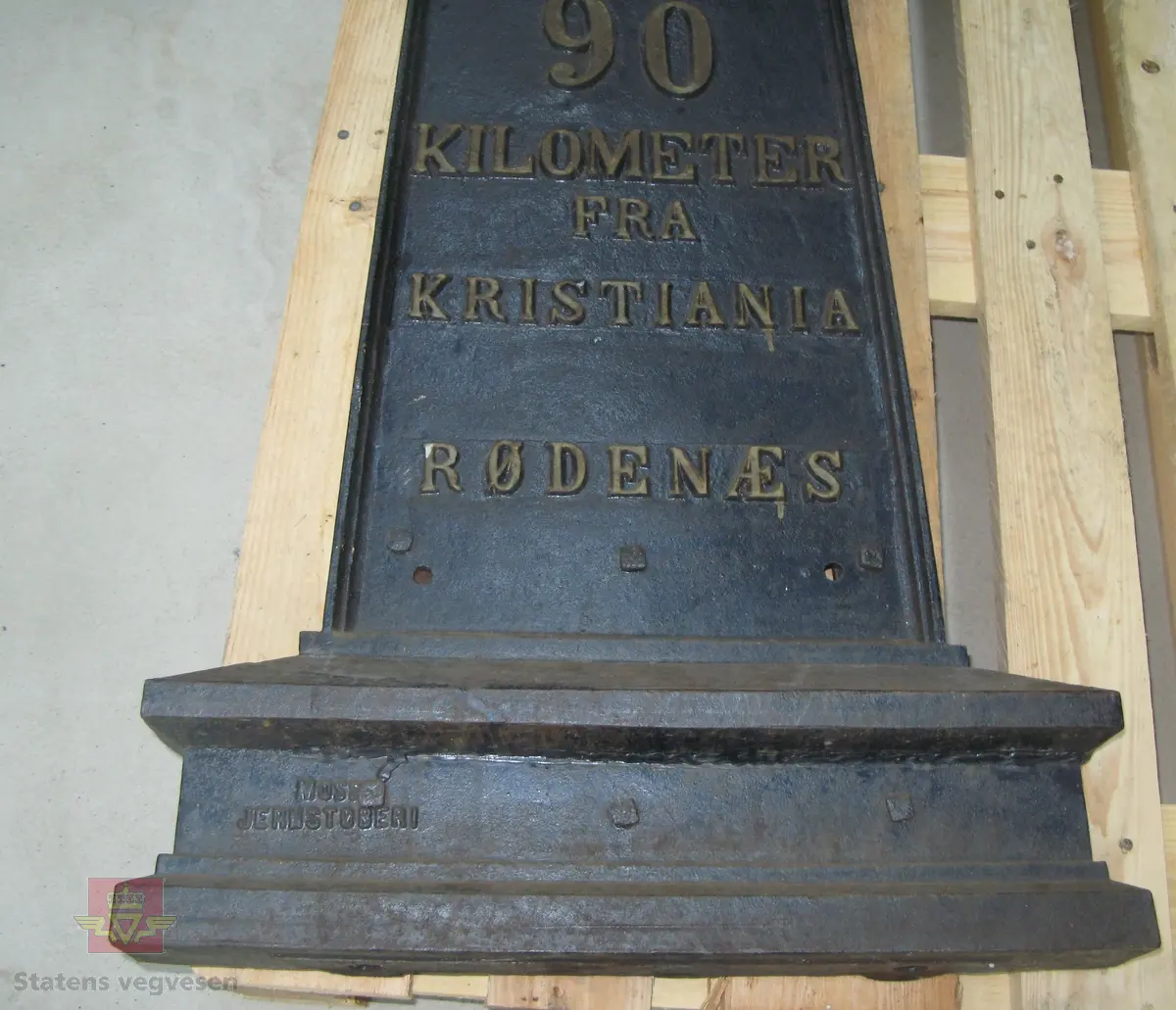 Kilometertavle, (skilt), lagd av støpejern, med riksvåpenmotiv og teksten:" SMAALENENES AMT 90 KILOMETER FRA KRISTIANIA RØDENÆS". Svart bunnfarge, tekst i gull. Baksiden har teksten: "MOSS JERNSTØBERI".