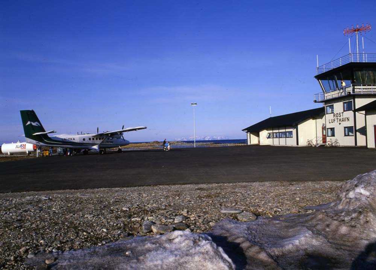 Lufthavn/Flyplass. Røst. Et fly, LN-FKA, DHC-6 Twin Otter fra Widerøe står parkert ved Ekspedisjonen/Flytårnet. I bakgrunnen fjell på Værøy.