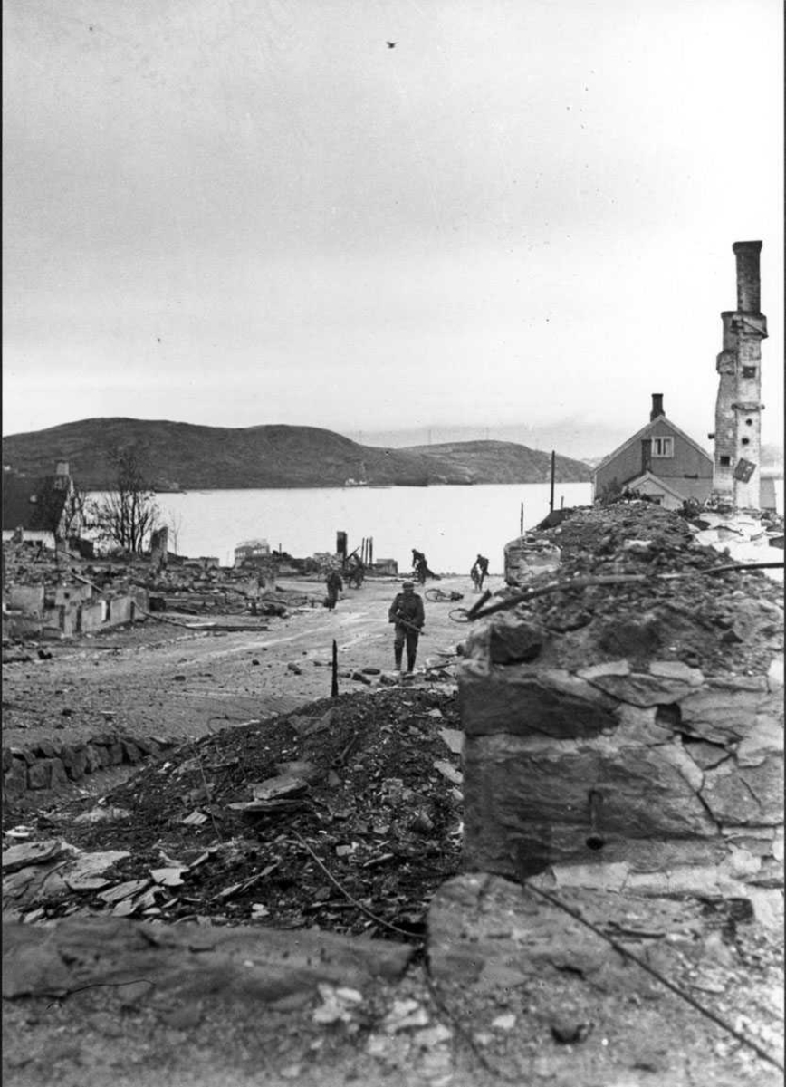 By - tettsted. Vei med noen personer, menn i militæruniform. Ruiner av flere bygninger. Hav - havneområde i bakgrunnen. Tyske soldater, hjertøya i bakgrunn.