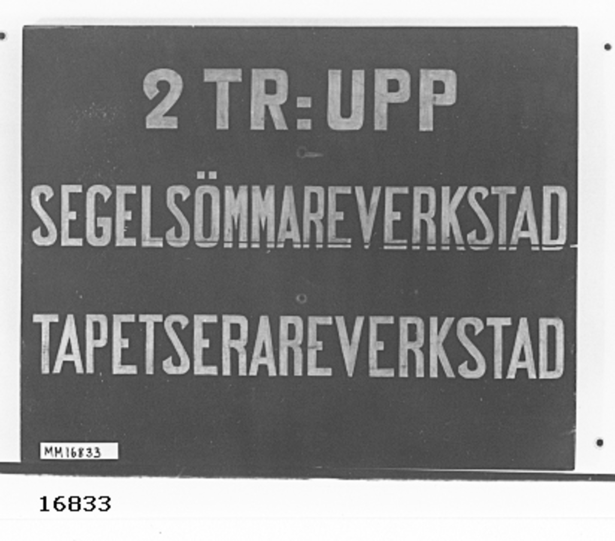 Skylt tillverkad av två sammanfogade furubrädor, målad i svart med vit text "2 TR:UPP SEGELSÖMMAREVERKSTAD TAPETSERAREVERKSTAD". Bokstävernas höjd 60 mm. I tavlans mitt två borrade hål för upphängning.