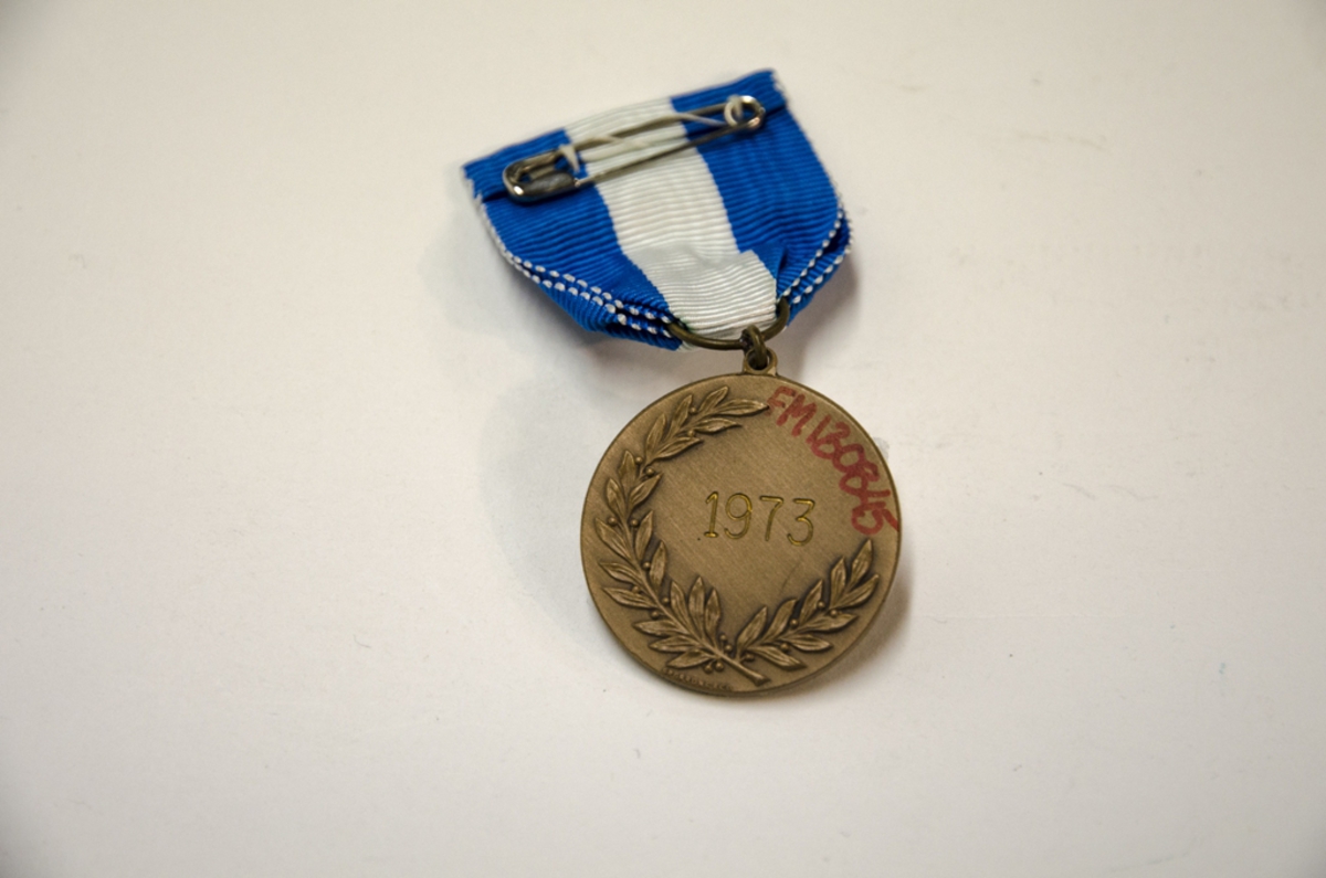 Idrottsmedalj i metall med motiv: Skidåkare. Medaljen har ett blå, vitt band och kommer från Nordenskiöldsloppet. På baksidan står ingraverat 1973.