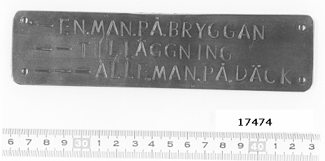 Rektangulär mässingsskylt med hål för upphängning i hörnen. Tre rader stansad text med signal framför:"-EN.MAN.PÅ.BRYGGAN.", "- - TILLÄGGNING" och "- - - ALLE.MAN.PÅ.DÄCK.".